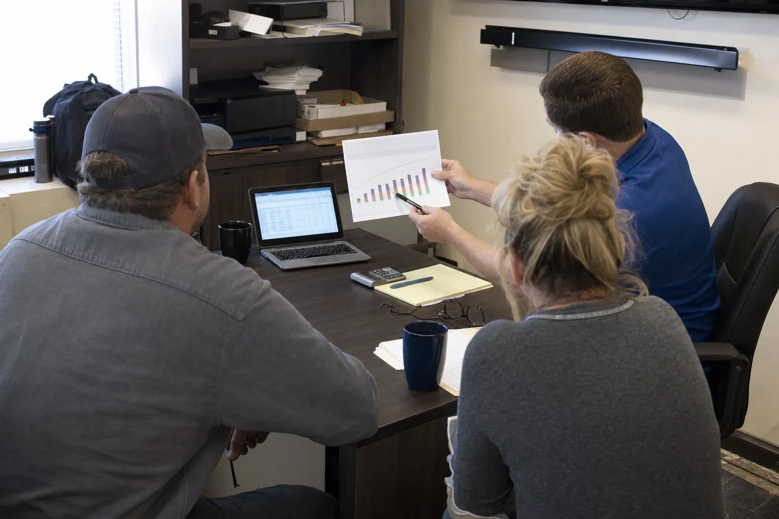 Agent in a blue polo holds up a printed bar chart for a man in a cap and a blonde woman seated at a desk in a small back office with a laptop and notepad